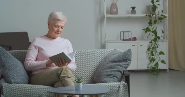 Dromerige senior ontspannen middelbare leeftijd volwassen Kaukasische vrouw draagt casual kleding lezen favoriete boek roman in grijze omslag draait pagina zitten op de bank het weekend doorbrengen in de woonkamer tijdens lockdown — Stockvideo