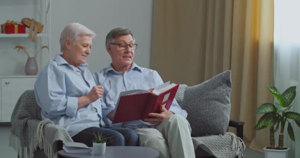 Gamla par kaukasiska äldre familj gråhårig man och kvinna morföräldrar sitter på soffan i mysigt vardagsrum tittar i fotoalbum minns trevliga stunder i livet njuter av minnen, nostalgi — Stockvideo