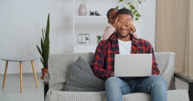 Afro-Amerikalı adam evinde oturuyor, online oyun oynuyor, bilgisayarında daktilo yazıyor, uzaktan çalışıyor, küçük zararlı kız odada gürültü yapıyor, babasının gözlerini kapatıyor, elleriyle oynamak istiyor.
