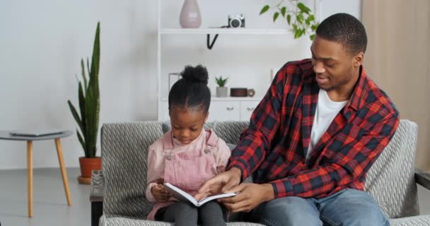 Afro-americano pai e bonito engraçado inteligente filha estudo juntos em casa leitura cinza livro virar página desfrutar romance conto de fadas pai solicita pouco pré-escolar menina sentado no sofá juntos casa educação — Vídeo de Stock