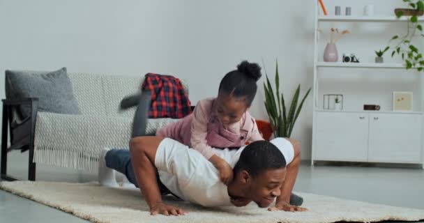 Un père célibataire souriant à la peau foncée faisant des pompes par terre avec une jolie fille d'âge préscolaire allongée sur le dos. African American papa s'amuser à la maison séance d'entraînement avec fille enfant bien-aimée, loisirs en famille — Video