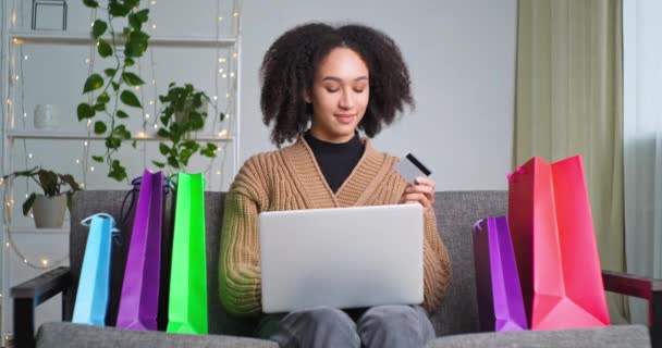 Hermosa mujer afroamericana feliz sentada en el sofá en casa recogiendo ropa nueva en la tienda en línea pedidos de entrega a través de compras en red utilizando el ordenador portátil y tarjeta de crédito se regocija con descuentos aplaude — Vídeos de Stock