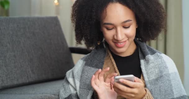 Retrato de menina sorridente com cachos afro mista senhora adolescente sentada em casa coberta com cobertor olha para tela do telefone mensagens de texto com namorado amado on-line em redes sociais alegra mensagem — Vídeo de Stock