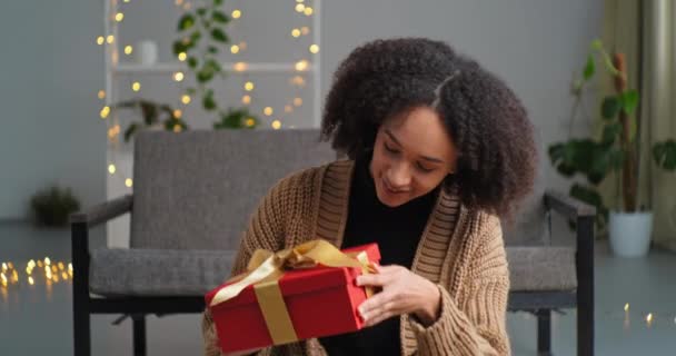 Vonzó elegáns afro-amerikai lány afro göndör nő hűvös frizura nevet ül a nappali padlóján nézi a kamera és a kezében piros ajándék dobozok rázza jelen furcsán érdeklődéssel — Stock videók