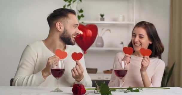 Pár szakállas fiatal kaukázusi férfi és barna lány őszintén nevetnek szórakozás együtt otthon Valentin nap évfordulója ünneplés alkalmazása a szemükre piros szív pózol a kamera — Stock videók