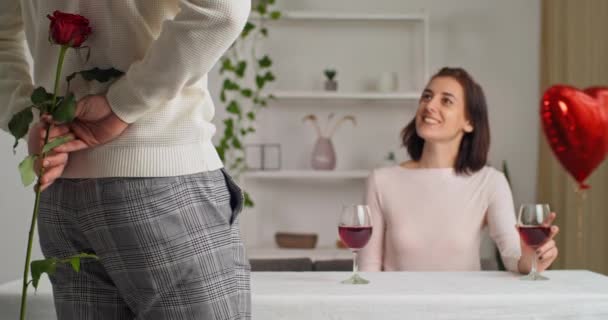 Вид сзади мужчина молодой красивый романтический парень с красной розой в руке делает его любимая жена девушка молодая женщина, сидящая за столом в кафе дома в ресторане дает цветок девушке — стоковое видео