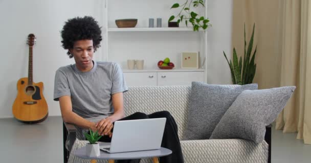 Un student afro-american serios care vorbeste pe telefonul mobil foloseste laptopul pentru a studia de la birou. Freelancer consultă prin smartphone comunică online în interiorul apartamentului — Videoclip de stoc