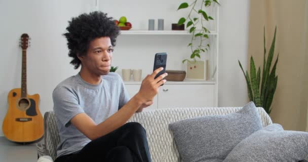 Emocionado africano adolescente estudiante ganador mantenga teléfono inteligente se siente sorprendido por la alegría con móvil apuesta en línea juego de la aplicación ganar sentado en el sofá de casa mirar el teléfono celular celebrar leer buenas noticias en las redes sociales — Vídeos de Stock