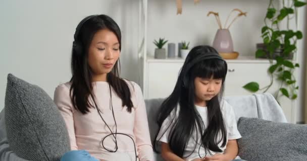 Азійська жінка-кореєць мати з маленькою дочкою, батьки тисячолітнього віку і етнічна дитина шкільного віку сидять разом на дивані, слухаючи пісні на навушниках, насолоджуючись мелодією і розслабленням. — стокове відео