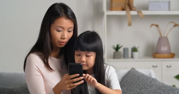 Calma focada mulher asiática jovem mãe solteira babá irmã mais velha sentado no sofá com menina filha criança olhando para a tela do smartphone móvel encomendar compras on-line usando o serviço on-line app — Vídeo de Stock