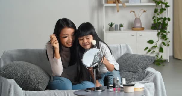 Little daughter and loving mother asian family two girls doing makeup beauty procedures at home sitting on couch looking in mirror use cosmetics mascara and blush take care of skin spend time together — Stock Video