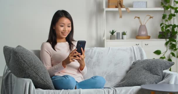 Asiática jovem mulher étnica sentada no sofá acolhedor na sala de estar usa telefone celular para se comunicar com os amigos faz chamada de vídeo remotamente de casa acenando saudação mão para webcam, conceito de tecnologia — Vídeo de Stock