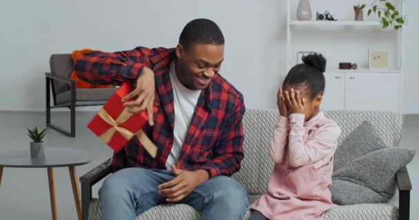 Kochający ojciec Afro amerykański mężczyzna siedzi na kanapie z córką zasłaniając jej oczy dłońmi tata robi nieoczekiwany prezent dla dziecka na jego urodziny daje czerwony prezent pudełko gratulacje przytula dziecko — Wideo stockowe