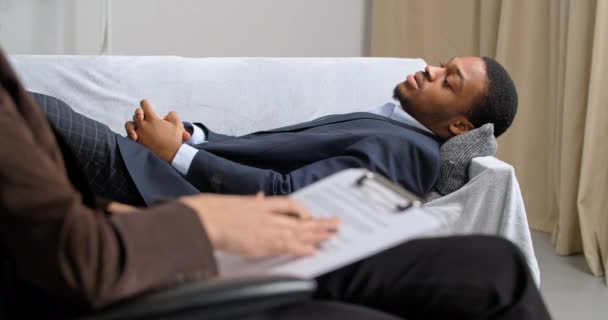 Afro homme d'affaires américain se trouve sur le canapé dans le bureau du psychologue consulte un psychiatre en cours psychothérapie de réadaptation parle avec spécialiste femme méconnaissable écrivant des notes dans un carnet — Video