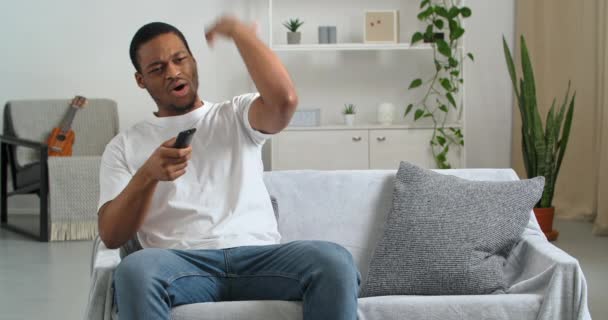 Afro americano cara fã emocional solteiro usa roupas casuais em casa sentado na sala de estar moderna torcendo por sua equipe favorita assistindo tv online transmitindo jogo de esportes feliz em ganhar sucesso — Vídeo de Stock