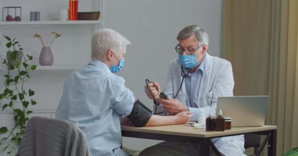 Két ember a klinikán, idősebb férfi orvos méri a betegek vérnyomását a rendszeres találkozón használ vérnyomásmérőt. Magas vérnyomásban szenvedő idős nő szakmai kardiológushoz megy — Stock videók