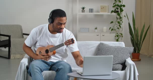 Hombre estudiante afroamericano usa auriculares aprendiendo nuevas habilidades durante cuarentena de encierro viendo lecciones de video en línea practicando jugar con un pequeño instrumento musical ukelele hobby ocupado creando canción — Vídeo de stock