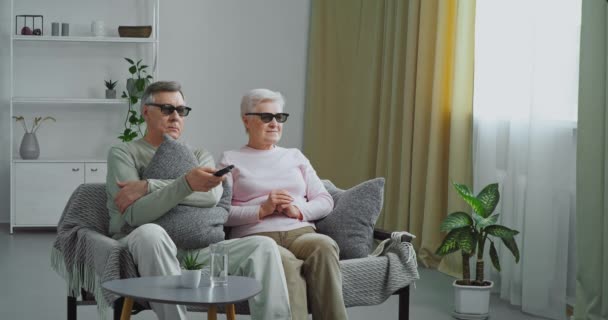 Серьезные ориентированные пожилая пара бабушка и дедушка старики носят трехмерные 3D очки, сидя на диване человек использует пульт дистанционного управления, чтобы переключить каналы, ищущие кино, домашний кинотеатр во время карантина — стоковое видео