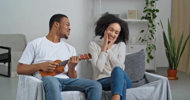 Romantický afro americký přítel sedí na gauči doma se svou milovanou přítelkyní během izolace v karanténě hraje ukulele malé kytary zpívá píseň serináda překvapí svou ženu s hudebním dárkem — Stock video