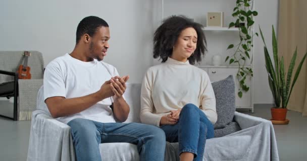 Афро-американская пара молодоженов бойфренд и подружка сидят дома диван ссорясь клянется ревнивый муж кричит женщина закрывая уши пальцами требует молчания делает запрещенный жест рукой — стоковое видео
