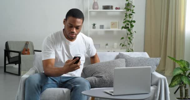 Afro american guy sleeping at home on couch on pillow waiting for call businessman freelancer wakes up from sound of mobile phone speaks on smartphone receives urgent information types data in laptop — Stock Video