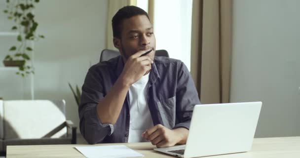 Pensive afro-amerikai diák srác töprengő ötlet jegyzeteket készít a jövőbeli projekt könyv egy papírlap tanulmány tárgya belenéz laptop képernyő ül asztalnál otthon vagy irodatervezés — Stock videók