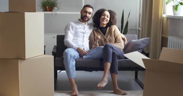 Gelukkig multi-etnisch paar zitten op de bank in de buurt van hoop bezittingen in kartonnen dozen, afro-Amerikaanse vrouw tonen aan de camera sleutels, symbool van purhapse van thuis. Opgewonden huiseigenaren portret, landgoed concept — Stockvideo