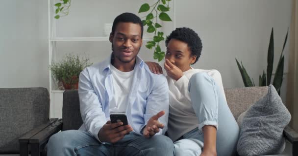 Afro-americano casal homem e mulher amigos sentados juntos no sofá na sala de estar interior olhando para a tela do telefone falando na câmera web respondendo videochamada discutindo rindo rindo assistindo vídeo online — Vídeo de Stock