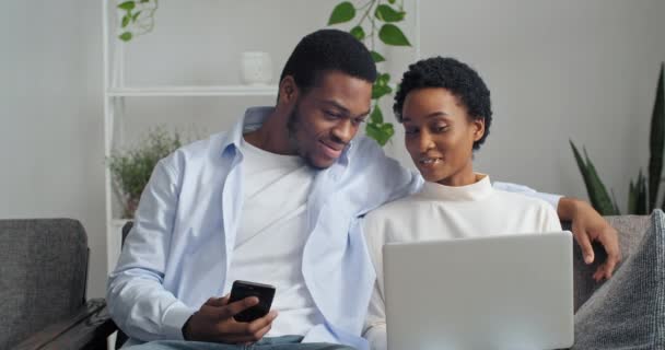Afro amerykański etniczny para młody rodzina czarny mężczyzna z telefonu i etnicznej kobiety z laptopem siedzi razem na kanapie w domu pokój dzienny czat rozmowy z przyjaciółmi odpoczynku pracy zdalnie — Wideo stockowe