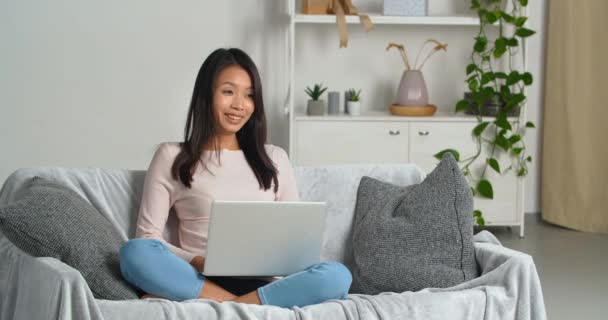 긍정적 인 만족을 느끼는 동양인 여성 이 거실에 편안 한 현대 소파에 앉아 있다. 노트북 서핑을 이용하고 농담 영화를 통해 진심으로 큰 웃음을 짓는다. — 비디오