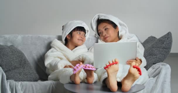 家庭温泉，亚洲母亲坐在舒适的沙发上，小女儿穿着浴衣和毛巾在洗澡后休息，用手提电脑孩子摸妈妈的鼻子来修脚 — 图库视频影像