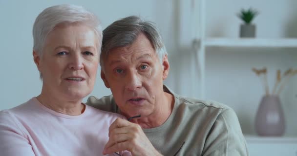 Elderly lively emotional couple sitting on couch records video message grey haired man points with finger direction to camera senior woman wave hands talking chatting using webcam during lockdown — Stock Video