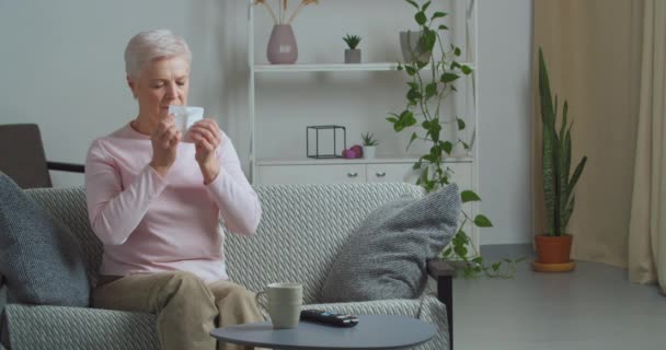 Kranke alte Frau Oma ältere Dame mit saisonaler Allergie Atemwegserkrankungen Symptome laufende Nase ausgeblasene Tücher mit Papierserviette sitzt auf Sofa im Wohnzimmer mit Tasse heißen Tee Medizin — Stockvideo