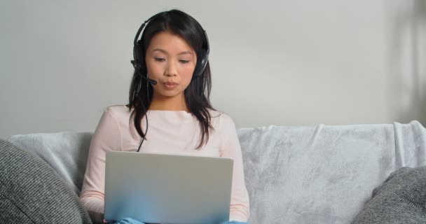 Konzentrierte junge asiatische Geschäftsfrau trägt ein Headset, das per Telefonkonferenz kommuniziert, spricht mit Blick auf den Laptop, der auf dem Sofa sitzt, Videochat-Bewerbungsgespräch oder Fernsprachkurs mit Online-Lehrer — Stockvideo