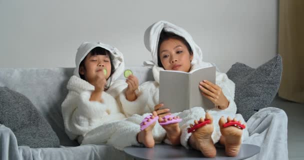 Две азиатки разных поколений в халатах, мать и дочь расслабиться после душа на диване чтении книги ребенок ставит свежие круги огурца на глаза делает натуральную маску едят овощи — стоковое видео