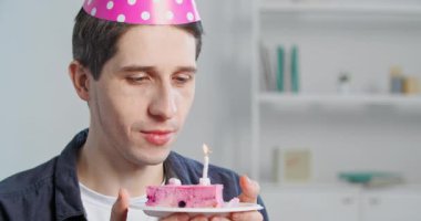 Bin yıllık mutlu beyaz adam portresi. Pembe parti şapkalı yalnız yetişkin bir adam. Elinde mumlar söndürülüyor.