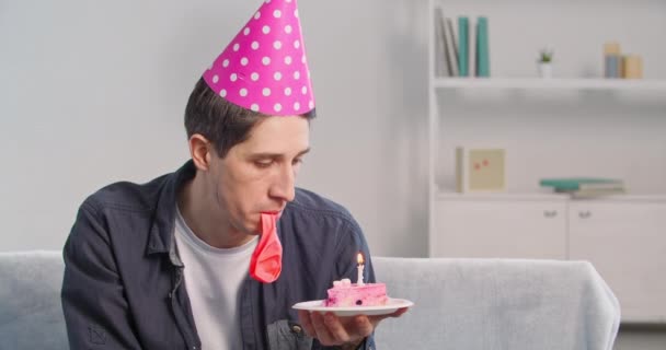 Portrnt single millennial man celebrating his birthday alone at home during quarantine wears pink hat on his head blowing up birthday candle on cake using balloon pretending happy feeling desperate — Stock Video