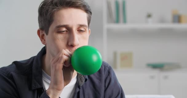 Close-up father man preparing for holiday birthday party, portrait of caucasian millennial guy doing effort inflates balloon inhales air into green rubber bubble looking at camera, celebration concept — Stock Video