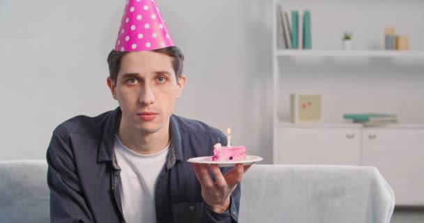 Ritratto di adulto caucasico giovane uomo triste seduto da solo a casa indossa festivo compleanno cappello rosa sulla testa tiene cupcake in mano pezzo di torta soffia candela rende desiderio si sente deluso tristezza — Video Stock
