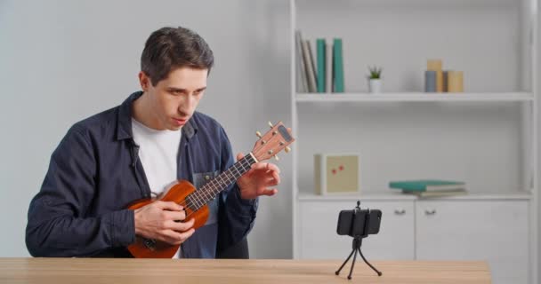 集中式高加索千禧年男教师在线导师的肖像演示了如何用智能手机记录音乐工作坊的对话来演奏小吉他ukulele在互联网课上的领唱 — 图库视频影像