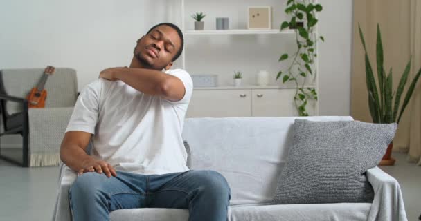 Афроамериканець, молодий етнічний хлопець у повсякденному одязі, сидить на дивані в незручному становищі, відчуваючи дискомфорт, коли біль у шиї страждає від хронічної хвороби, що масажує м'язи спини рукою. — стокове відео