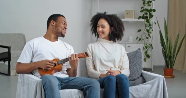 Молодой любящий афро-американец парень муж поет песню, играя на маленьком музыкальном инструменте укулеле для любимой красивой женщины кудрявая жена подружка делает творческий подарок сидя дома диван — стоковое видео