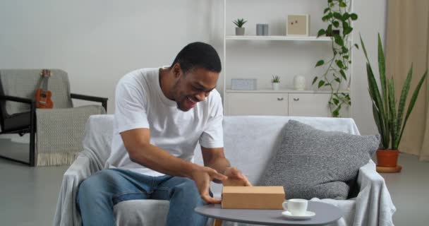 Zvědavý africký americký spotřebitel příjemce sedí doma obdrží balíček s dárkem z internetového obchodu tře si ruce se zájmem rozbaluje kartónové krabice drží šedý notepad raduje v překvapení — Stock video