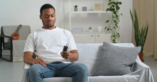 Afro amerikansk ung kille ungkarl tusenårig svart man sitter på soffan hemma titta på tv använder fjärrkontroll för att växla kanaler letar efter favorit sportprogram eller film, helg koppla av koncept — Stockvideo