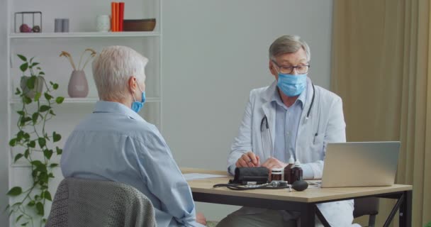 高齢の医師の専門家は、医療マスクコンサルタントを身に着けていますシニア女性患者は、紙の上に処方を書きますクリニックでの会議で慢性疾患の話のための薬のボトルを示しています — ストック動画