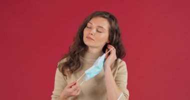 Kafkasyalı genç, sorumluluk sahibi güzel bir kız mavi stiryl tıbbi maske takıyor. Başparmaklarını kaldırıyor. Stüdyoda kırmızı arka planda poz veren önlemlere olumlu bir tavır sergiliyor.