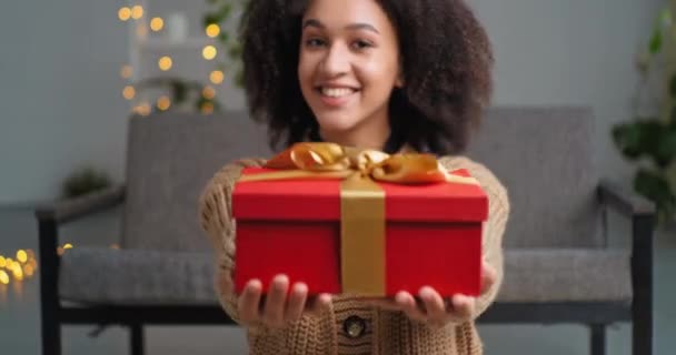Όμορφη γενναιόδωρη αφροαμερικανή γυναίκα εθνοτική κορίτσι με κόκκινο δώρο κουτί χαμόγελα ειλικρινά κάθεται στο ζεστό σπίτι σαλόνι πάτωμα συγχαίρει για τις διακοπές κοιτάζοντας κάμερα, εορταστική έννοια κόμμα — Αρχείο Βίντεο