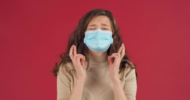 Chica joven con el pelo rizado lleva máscara médica protectora gira los dedos rezando con la esperanza de éxito pide destino y dios para buenos resultados de prueba para coronavirus se encuentra aislado en el fondo rojo en el estudio — Vídeos de Stock