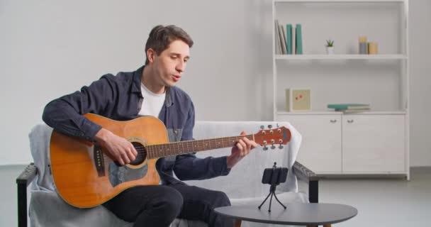 Emberek és művészetek, kaukázusi Y-generációs férfi ül kényelmes otthoni felvétel videó online hívás internetes gitár lecke tanítás zenét távolról beszél a telefon kamera küld légcsókok — Stock videók