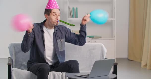 Веселий активний кавказький чоловік вдома під час блокування робить відеодзвінок привітання подрузі на день народження дистанційно, використовуючи відеодзвінки, що махають кульками, носить святковий капелюх, що дме, створює шум — стокове відео